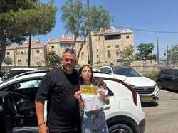 תלמידה מציגה שלט נהג חדש ליד רכב ואבי